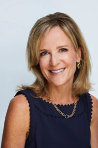 Smiling woman with blonde hair wearing a navy top and gold necklace against a light background